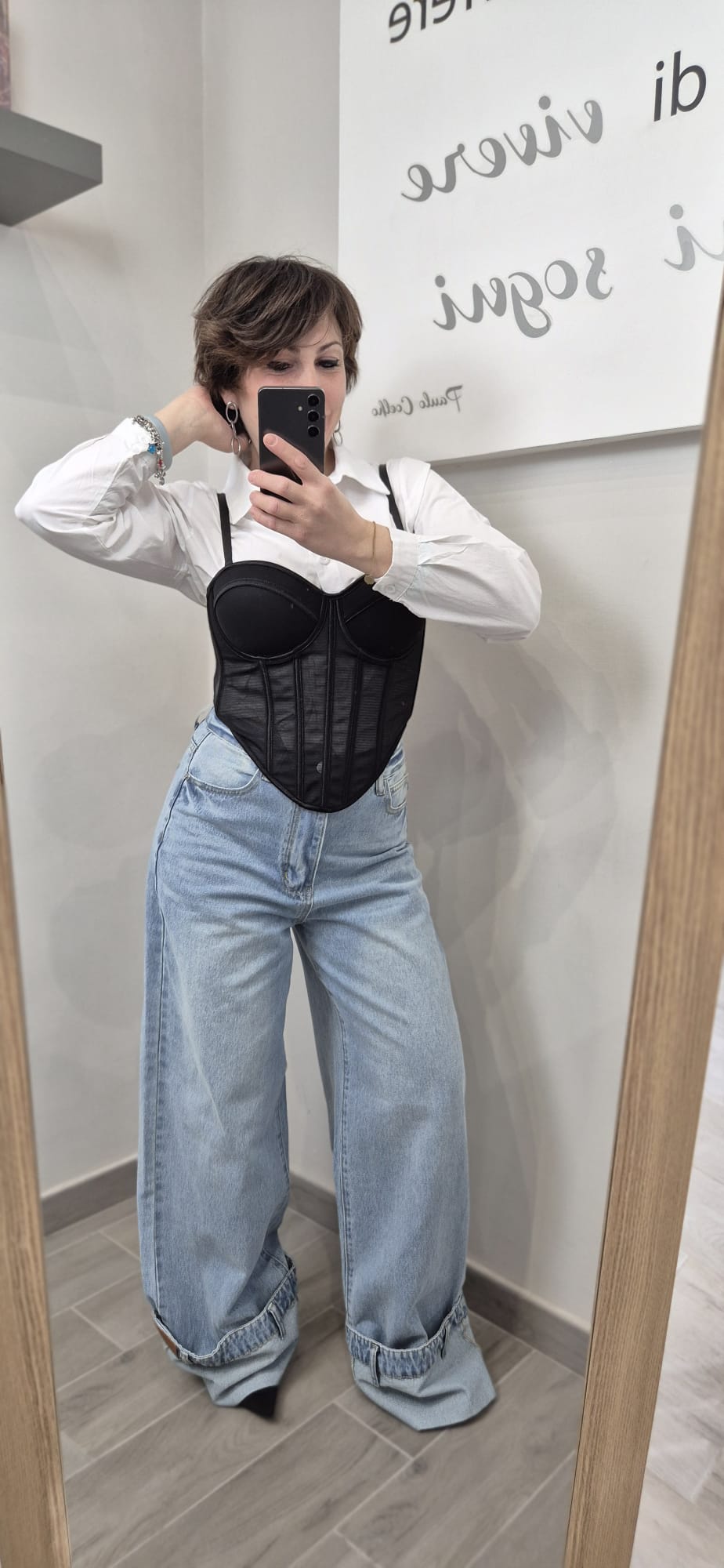 Donna indossa camicia bianca, corsetto nero e jeans larghi a vita alta davanti allo specchio.