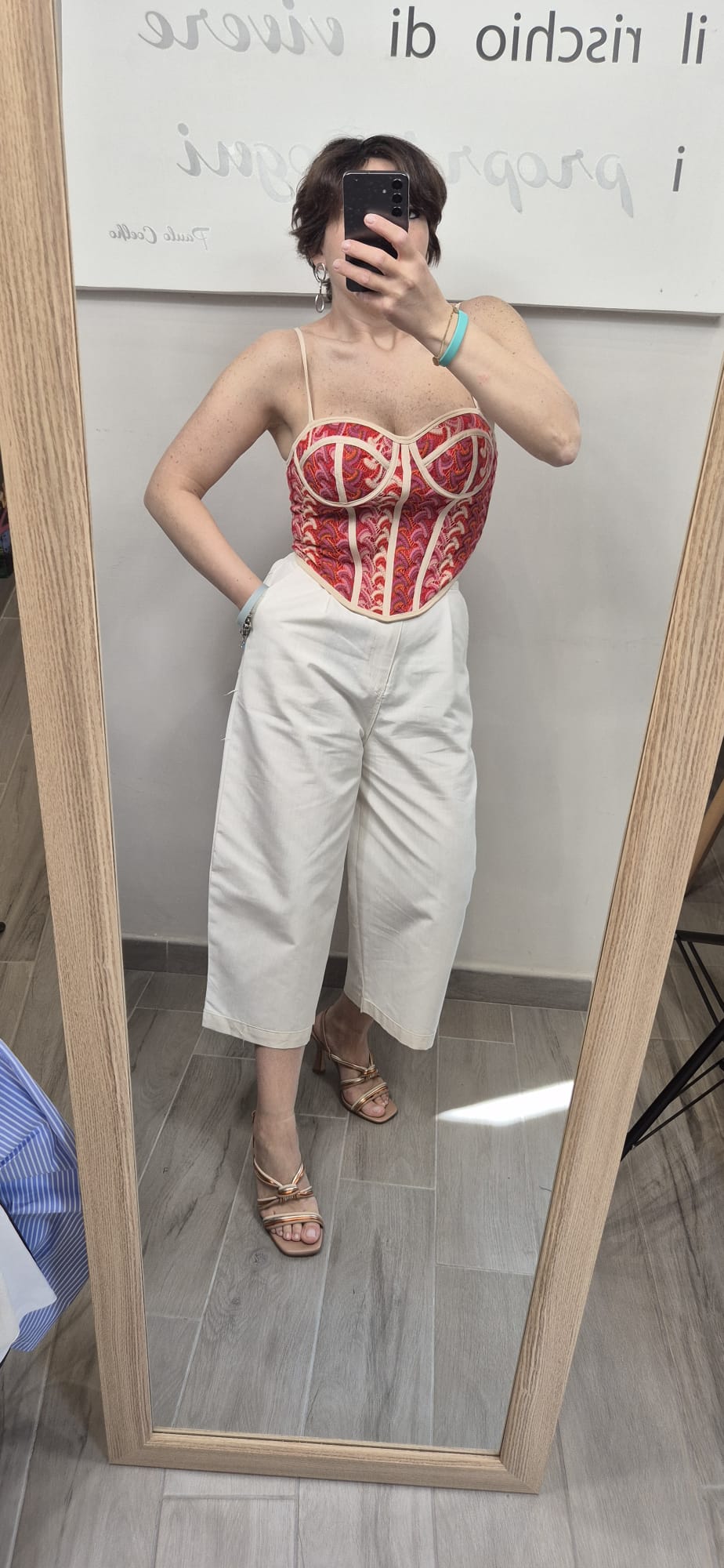 Donna con top bustier rosso fantasia e pantaloni bianchi a palazzo, sandali oro, selfie allo specchio.